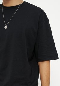 Άτομο που φοράει απλό μαύρο κοντομάνικο t-shirt και ασημένιο κολιέ με στρογγυλό μενταγιόν, πάνω σε λευκό φόντο.