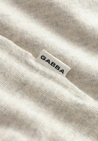 Ελαφρώς μπεζ ύφασμα με απαλή υφή, με ορατή ετικέτα που γράφει "GABBA" με μαύρα γράμματα. Οι ίνες παρουσιάζουν μικρές διαφορές χρωμάτων.