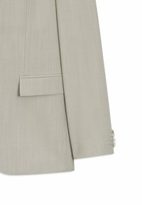 Manche et taille d'un blazer beige clair avec poche à rabat et quatre petits boutons sur le poignet, présenté sur fond blanc.