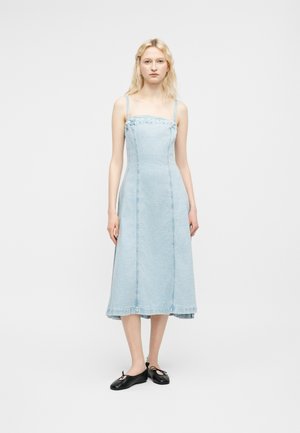 Noor naine, kellel on pikk blond juuksed, seljas helesinine teksast ilma varrukateta midi kleit ja mustad baleriinikingad, seisab valgel taustal.