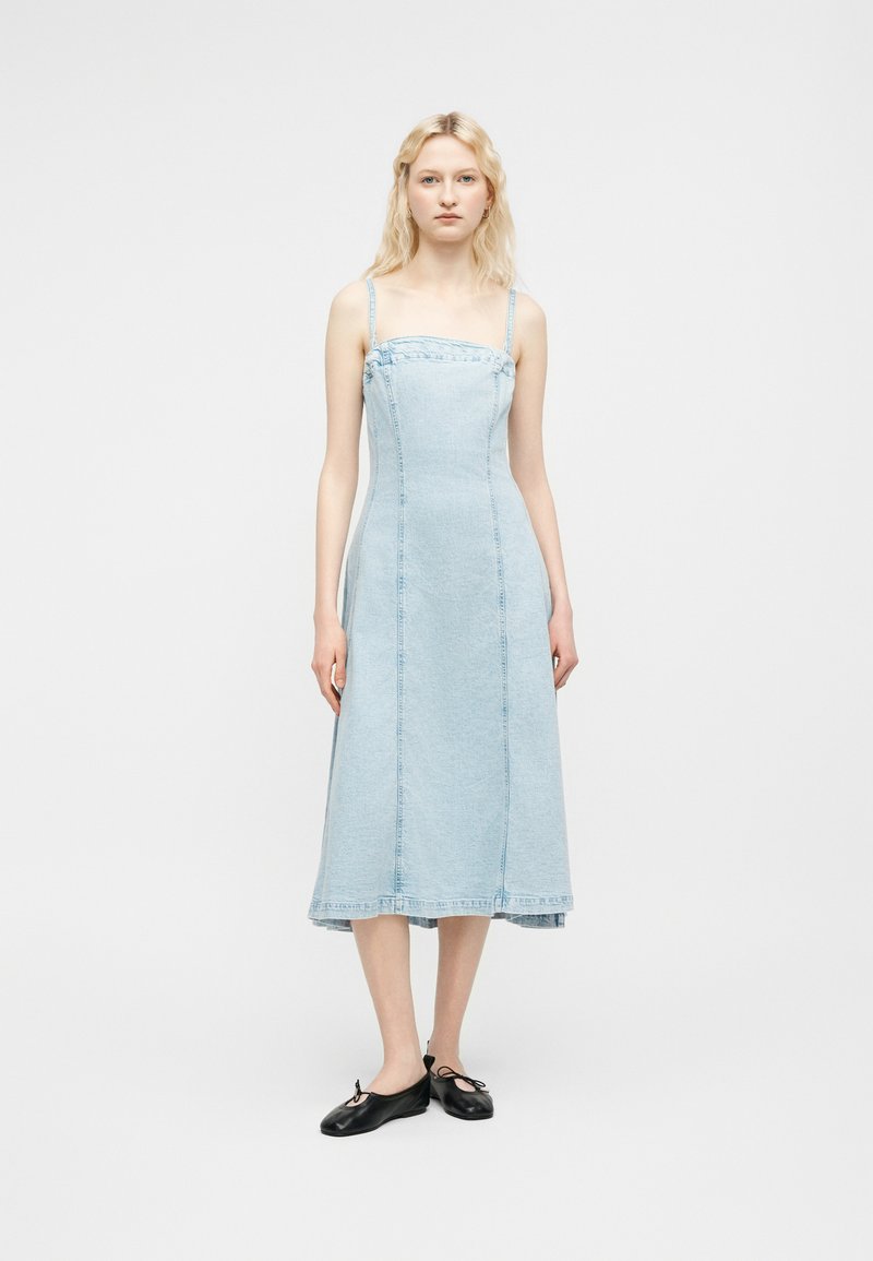 Νεαρή γυναίκα με μακριά ξανθά μαλλιά που φοράει αμάνικο μίντι φόρεμα από ανοιχτό μπλε τζιν και μαύρα μπαλέτα, που στέκεται μπροστά σε λευκό φόντο.