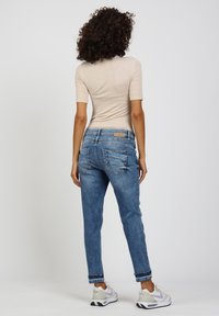 Frau mit lockigem Haar trägt ein beiges Kurzarmoberteil, blaue Jeans mit umgeschlagenen Säumen und weiße Turnschuhe, steht vor einem einfarbigen Hintergrund.