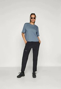 Sinine lühikeste varrukatega t-särk väikese logoga, koos mustade pükstega, millel on lukuga taskud, ja mustad sportlikud jalanõud.