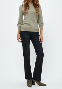 Kvinna i olivgrön tröja, svarta raka jeans och olivgröna mockaskor med blockklackar, stående mot en neutral bakgrund.