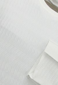 Witte textuurstof met een golvend patroon, voorzien van een vierkante zak. Het materiaal lijkt lichtgewicht en enigszins geribbeld.