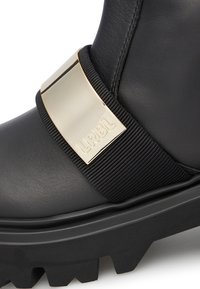 Black leather boot featuring a prominent gold metal strap with engraved "LRBL," textured rubber sole, and a chunky, rugged design.