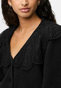 Melnā džemperis ar teksturētu audumu, mežģīņu apkakli ar viļņveida malām un atbilstošu melnu apdari. Poga aizdare priekša.