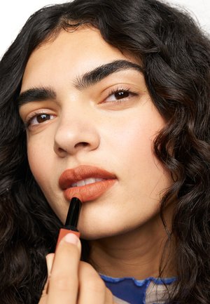 Matte brown lipstick is applied to lips; model features curly dark hair, clear skin, and defined brows, with a blue top visible.
