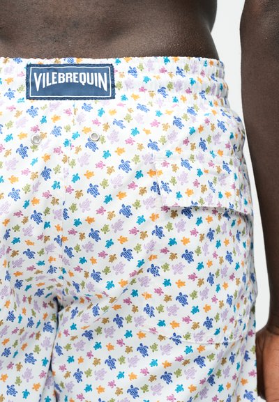 Κοντινό πλάνο λευκών μαγιό με πολύχρωμο σχέδιο χελωνών, με ετικέτα λογότυπου Vilebrequin και πίσω τσέπη σε άτομο με σκούρο δέρμα.