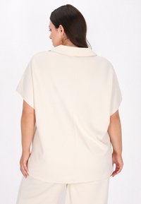 Ljust beige blus med ett texturerat geometriskt mönster, korta ärmar och en bred krage, med en lös och avslappnad passform och mjukt tyg.