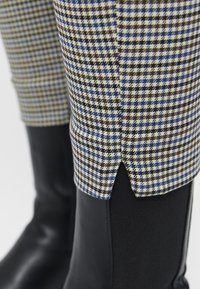 Gros plan sur un pantalon à motif pied-de-poule bleu et marron rentré dans des bottes en cuir noir avec des panneaux latéraux élastiques.
