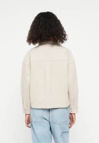 Light beige cropped jacket with a collar, featuring a back pleat and buttoned cuffs, paired with light blue denim trousers.