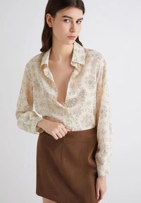 Blusa crema con motivi floreali marroni ondulati, design con bottoni e vestibilità morbida. Abbinata a una mini gonna marrone. Texture setosa.