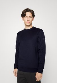 Sweatshirt bleu marine avec un col rond, des manches longues et des poignets côtelés. Présente un tissu texturé et un patch logo discret sur la manche gauche.