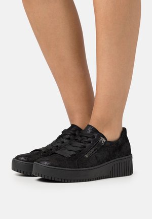 Jambes portant des baskets plateformes noires avec des lacets et des fermetures éclair latérales, présentées sur un fond clair uni.