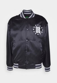 Chaqueta bomber de satén negra con cuello y puños acanalados en rayas grises, que presenta una letra "H" blanca y un patrón de estrellas en el lado izquierdo del pecho.