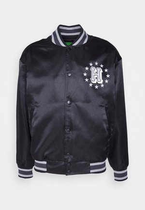 Chaqueta bomber de satén negra con cuello y puños acanalados en rayas grises, que presenta una letra "H" blanca y un patrón de estrellas en el lado izquierdo del pecho.