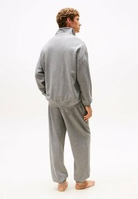Grijze katoenen blend trainingspak met een rits-hoogsluiter, losse pasvorm en elastische manchetten. Model staat met de rug naar de camera in een ontspannen houding.