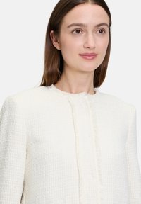 Biała teksturowana kurtka z postrzępionym kołnierzem, z wzorem wplecionym i dopasowanym krojem. Minimalistyczny design bez widocznych elementów metalowych.