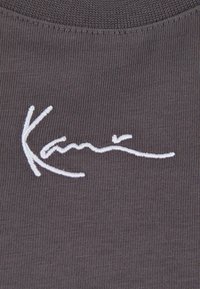 Harmaa kangasta, jossa on uritettu pinta, ja siinä on näkyvä valkoinen brodeerattu signeerauskuvio.