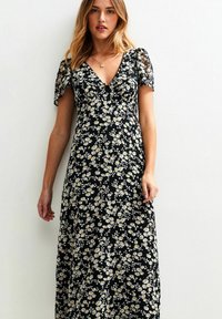 Mujer vistiendo un vestido midi negro con estampado floral en blanco y amarillo, con escote en V y mangas cortas abullonadas, de pie contra una pared blanca.