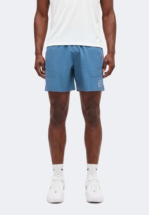 Mann trägt hellblaue Sportshorts, weiße Sneakersocken, weiße Sportschuhe und ein weißes Sportshirt und steht vor einem einfarbigen Hintergrund.