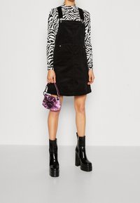 Svart korduroyklänning med justerbara axelband, kombinerad med en zebramönstrad långärmad topp. Accessoarer inkluderar en glänsande rosa handväska och svarta platåstövlar.