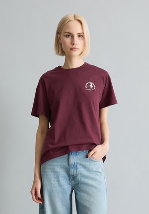 Blonde Frau mit kurzen Haaren, die ein burgunderrotes "Athletic Club" T-Shirt und hellblaue Jeans trägt, vor einem schlichten grauen Hintergrund stehend.