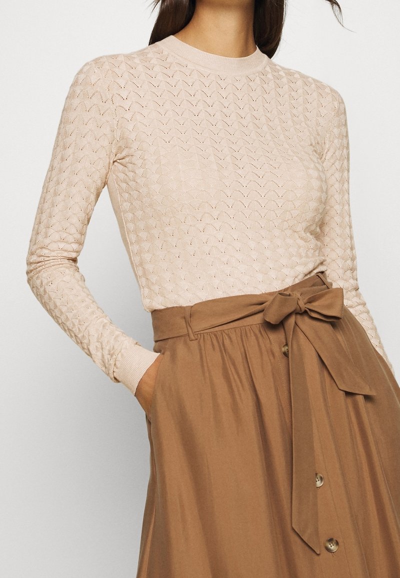 Femme portant un haut beige à manches longues à motifs, rentré dans une jupe marron taille haute avec une ceinture nouée et des boutons sur le devant.