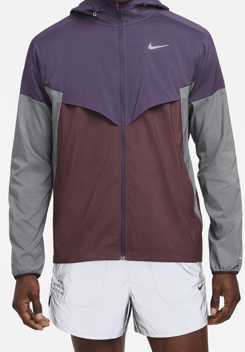 Homme portant une veste zippée Nike violette et grise avec capuche et un short blanc Nike, debout avec les bras détendus le long du corps.