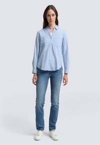 Light blue long-sleeve button-up shirt with two chest pockets, paired with fitted blue jeans and white sneakers, on a plain background.