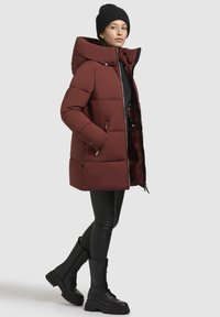 Rød pufferjakke med hætte, lynlås foran og sidelommer. Sorte leggings og snørebåndsstøvler i sort fuldender outfittet.