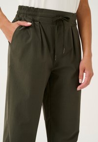 Pantaloni a sigaretta verde oliva realizzati in tessuto liscio. Presentano una vita elasticizzata, chiusura con cordoncino e tasche laterali. Dettaglio plissettato nella parte anteriore.