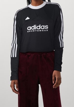 Person iført en sort Adidas langærmet trøje med hvide striber og logo, kombineret med mørkerøde fløjlsbukser mod en ensfarvet baggrund.