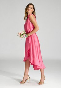 Robe en satin rose avec un ourlet voluminous, un col montant et une coupe décontractée. Portée avec des talons dorés à lanières et tenant un bouquet de fleurs.