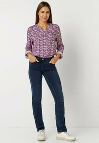 Langärmlige lila gemusterte Bluse mit V-Ausschnitt, kombiniert mit dunkelblauen eng geschnittenen Jeans und weißen Sneakers. Lässiges, bequemes Outfit.