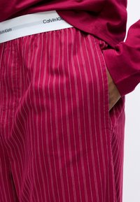 Pánské pyžamové kalhoty Calvin Klein v tmavě růžové barvě, s tenkými bílými vertikálními pruhy, elastickým pasem a bočními kapsami.