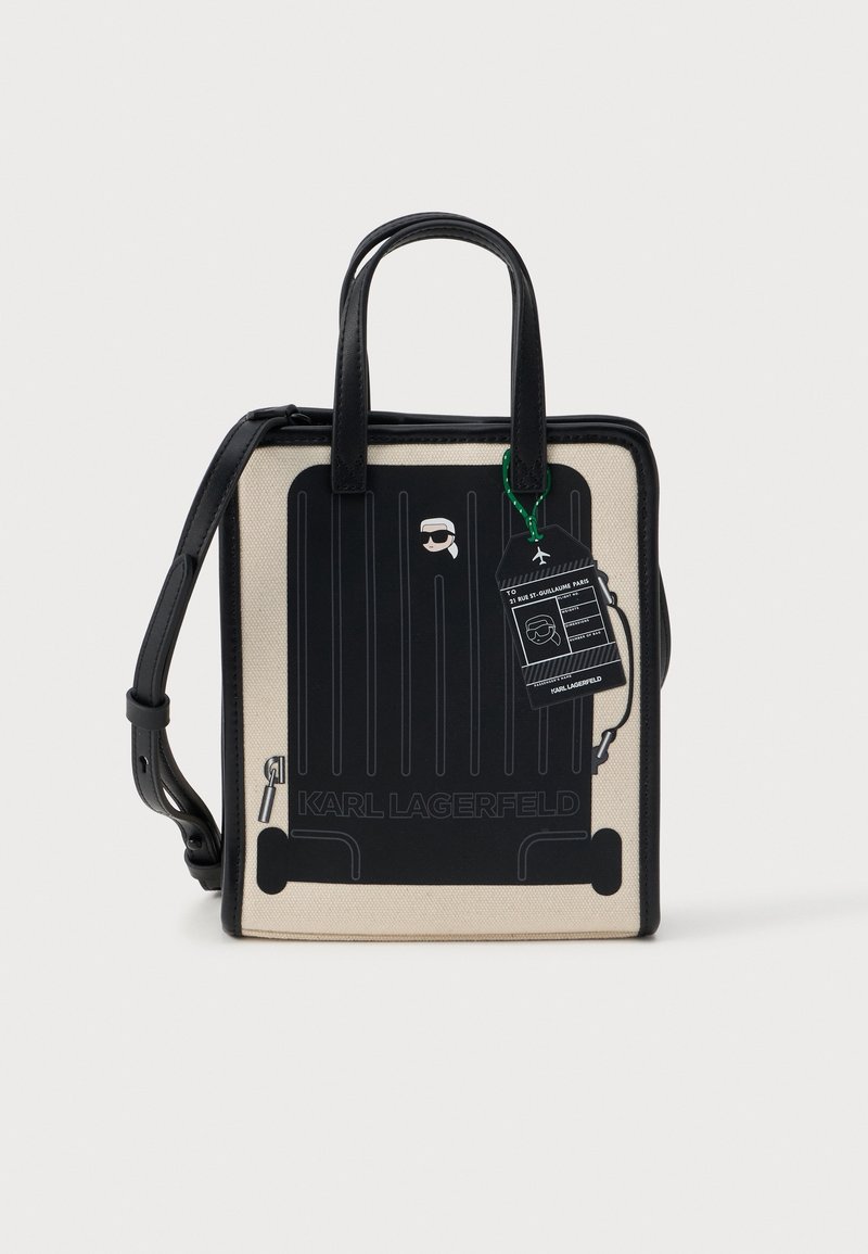 Sac à main carré beige et noir avec poignées noires, motif de valise et étiquette de bagage attachée avec un visage stylisé portant des lunettes de soleil.