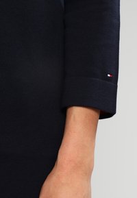 Marinblå stickad tröja med ärm som har en rullad manschett; har en liten röd, vit och blå logotyp vid ärmslutet. Mjuk textur.