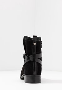 Stivaletto in suede nero con cinturini in pelle, zip sul retro e tacco basso impilato su sfondo bianco in studio.