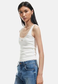 Witte geribde tanktop met brede schouderbanden en een aansluitende pasvorm, gepresenteerd met blauwe versleten denim jeans. Opvallend geborduurd logo aan de voorkant.