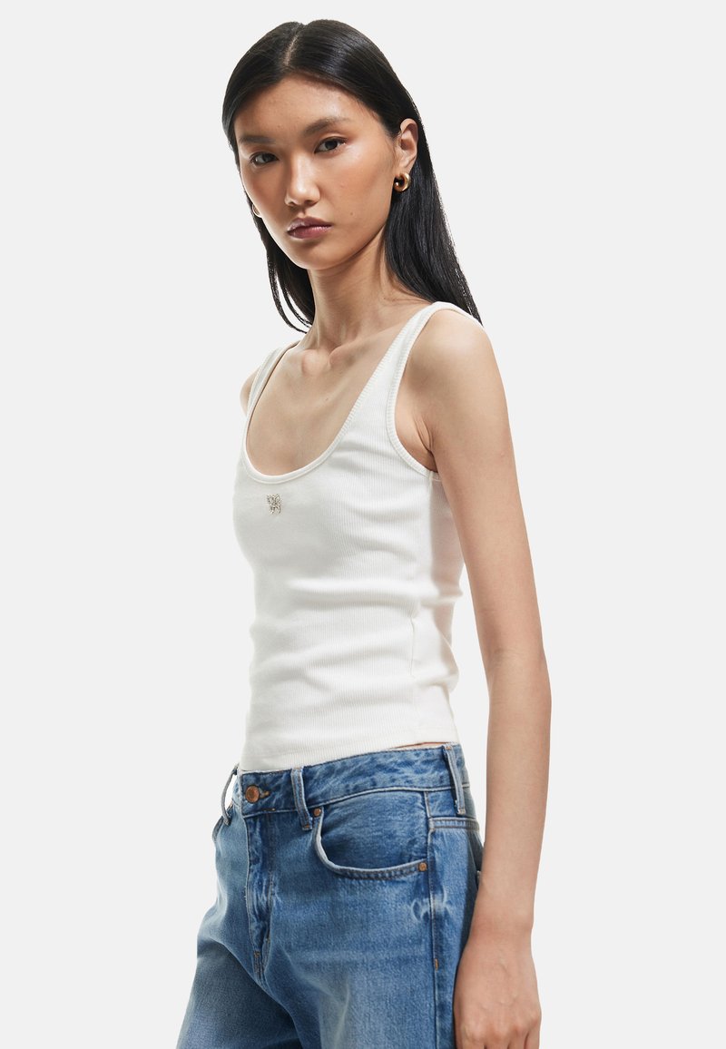 Witte geribde tanktop met brede schouderbanden en een aansluitende pasvorm, gepresenteerd met blauwe versleten denim jeans. Opvallend geborduurd logo aan de voorkant.
