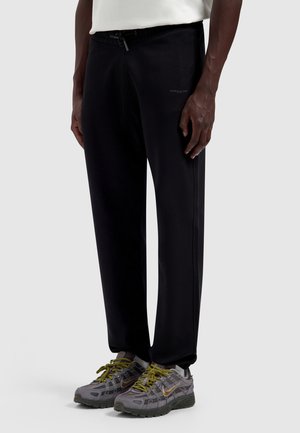 Man draagt zwarte insteekbroek en grijze sportschoenen met olijfgroene veters, staand tegen een effen lichte achtergrond.