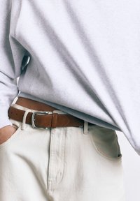 Person iført lysebeige jeans og en lysegrå sweatshirt med et brunt læderbælte og den ene hånd i lommen.