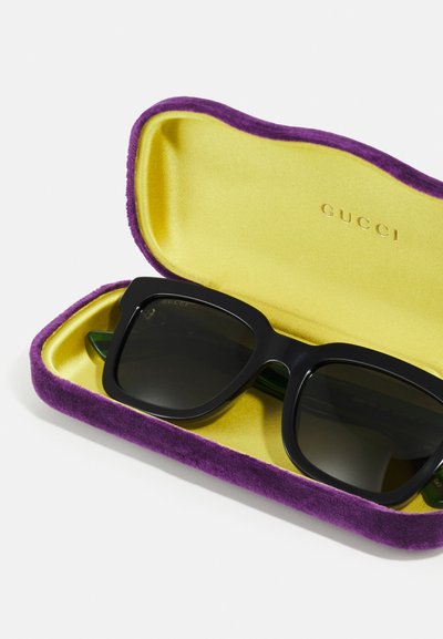 Occhiali da sole Gucci con montatura nera e lenti scure, riposti in un astuccio foderato in velluto giallo con esterno viola e logo dorato all'interno.