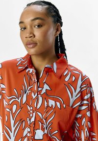 Camisa de patrón floral rojo con cuello, cierre de botones en la parte delantera y un bolsillo, hecha de una tela suave con acentos en blanco y negro.