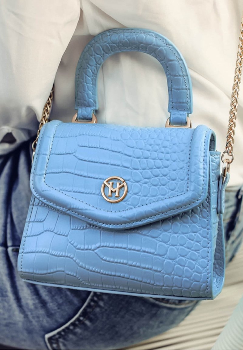 Petit sac à main bleu avec texture crocodile et bandoulière chaîne dorée porté par une personne en haut blanc et jean bleu.