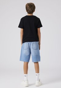 Črna bombažna majica, svetlo modri denim kratki hlače, bele športne copate in bele nogavice z črnim napisom, prikazano s hrbtne strani.