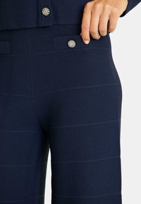 Pantalons texturés bleu marine avec un motif à carreaux et des boutons décoratifs en argent sur les poches, avec une finition lisse et une coupe décontractée.