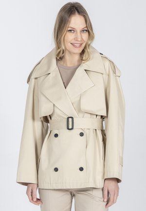 Kobieta o blond włosach, ubrana w beżowy płaszcz trencz z paskiem, na jasnych spodniach, uśmiechająca się na jednolitym szarym tle.